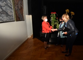 Dwie kobiety w maseczkach ochronnych gratulują sobie, a jedna z nich wręcza bukiet czerwonych róż drugiej. W tle czarna kotara. Po lewej stronie wisi obraz na ścianie.