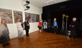 Uczestniczki wystawy stoją pod ścianą, na której wiszą obrazy. W tle widoczna czarna kotara.