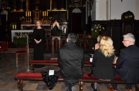 Kobieta w eleganckiej, czarnej, długiej sukni śpiewa. Przed nią znajduje się publiczność siedząca na ławkach kościelnych. W tle widać ołtarz. W kościele są zgaszone światła.