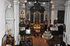 Białe ściany kościoła wraz z kontrastującym ciemnym ołtarzem. Zdjęcie zostało zrobione z perspektywy chóru kościelnego. Na dole widać ławki zasiadane przez ludzi.