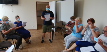 Warsztaty teatralne - zajęcia praktyczne dla uczestników. Każdy z uczetników ma założoną maskę na twrzy. Mężczyzna trzyma kartkę w rękach i przemawia do uczestników.