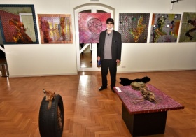 Andrzej Dziubek w czapce i okularach stoi obok prac wystawowych. Na fotografii widać lisa w oponie i szkielet zwierzęcia.