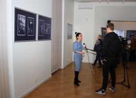 Edyta Pilichowska nagrywa swoje wystąpienie. Młody chłopak jest operatorem kamery. W tle widać fotografie wiszące na ścianie.