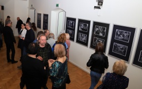 Uczestnicy wystawy dyskutują i podziwiają fotografie wiszące na ścianach.  Panuje gwar, wszyscy skupiają się na innej fotografii.