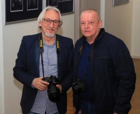 Dwóch mężczyzn z aparatkami fotofraficznymi pozuje do zdjęcia. Jedna lustrzanka jest z firmy NIKON.