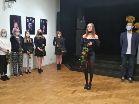 Na środku przed czarną kotarą i głośnikiem stoi młoda kobieta z czerwoną różą w ręce. Obok sceny po lewej stronie w oddali stoją trzy inne kobiety z kwiatami w ręce. Na ścianie wiszą dwie fotografie.