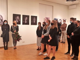 Młode kobiety w maseczkach ochronnych stoją na środku sali. Z tyłu ich można dotrzeć innych uczestników. Po lewej stronie widać kobietę trzymającą bukiet czerwonych róż. Na ścianie wiszą fotografie.