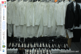 Białe ubrania wiszące na wieszaku. Na dole czarne szpilki ułożone w dwóch rzędach. Po lewej stronie logo organizatorów.