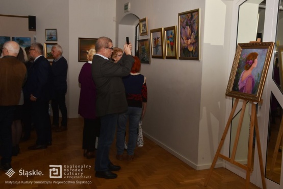 Grupa osób oglądających wystawę. Jeden z mężczyzn robi zdjęcie obrazowi przed nim wiszącym. Na ścianie wiszą obrazy a jeden z obrazów stoi na sztaludze. 