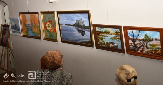 Dwie seniorki oglądające wystawę wiszących na ścianie obrazów. Obrazy przedstawiają pejzdaże i martwą naturę.